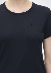 Gros plan d'une personne portant un t-shirt noir uni à col rond avec un petit logo noir brodé sur la poitrine gauche.