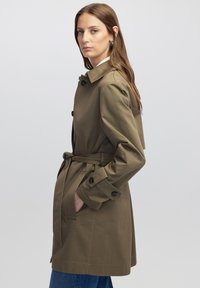 Abrigo trinchera verde oliva con cintura ceñida, frente abotonado y cuello ancho. Presenta un tejido texturizado y bolsillos laterales.