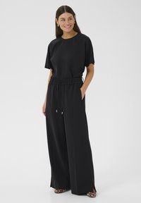 Čierny overal s uvoľneným strihom, elastickým pásom, krátkymi rukávmi a bočnými vreckami. Nosený s topánkami s leopardím vzorom, ktorý ukazuje minimalistický dizajn.