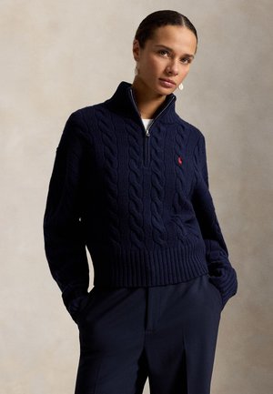 Kvinde iført en marineblå kabelstrikket sweater med kvartlynlås, ribkant nederst og på ærmerne, kombineret med mørke bukser, stående mod en neutral baggrund.