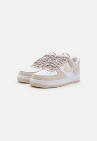 Le sneakers Nike Air Force 1 presentano una tomaia in pelle bianca con accenti in suede beige, punta rotonda, lacci piatti e una suola in gomma testurizzata.