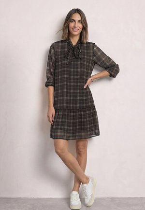 Femme aux cheveux mi-longs portant une robe à carreaux noire avec un nœud au col et des baskets blanches, debout contre un mur clair uni.