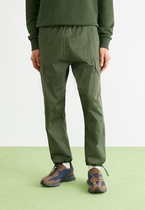 Groene cargobroek met een elastische tailleband, taps toelopende enkels en meerdere zijzakken. Draagt met bruine en marineblauwe sportschoenen.