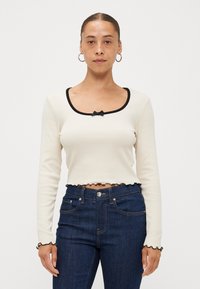 Cream long-sleeve top with black trim, bow detail at neckline, cropped length, and ruffled hem. Paired with dark blue high-waisted jeans.