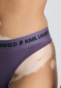 Fioletowe bawełniane majtki bikini z czarnym elastycznym pasem w talii, na którym znajduje się marka "KARL LAGERFELD" oraz sylwetka logo.