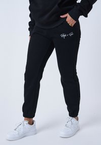 Pantalones de chándal negros con puños elásticos, con bordado blanco "Project x Paris" en el muslo izquierdo. Llevados con zapatillas blancas.
