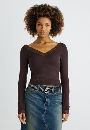Γυναίκα με σγουρά μαλλιά φοράει σκούρο καφέ μπλούζα λαιμόκοψη V με δαντελωτό τελείωμα και ζώνη με τρουκς πάνω από μπλε τζιν φούστα.