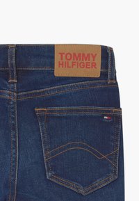 Pantalones vaqueros de mezclilla de color azul oscuro, con un parche de cuero marrón etiquetado "TOMMY HILFIGER", costuras decorativas y un bolsillo trasero.