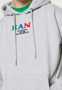 Felpa grigia con cappuccio e lacci, con testo colorato "KANI" e "THE ORIGINAL EST. 1989" in nero, realizzata in tessuto morbido.