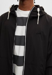 Svart zip-up jacka med en texturerad yta, vita snörningsdetaljer, bärs över en grå och vit randig t-shirt.