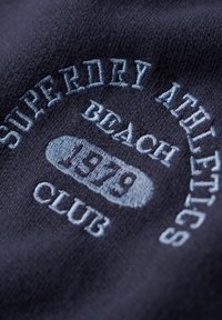 Tessuto di maglia blu navy con testo ricamato azzurro: "SUPERDRY ATHLETICS BEACH CLUB 1979." Texture liscia con un design circolare.