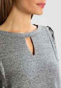 Szara bluzka z okrągłym dekoltem z wycięciem w kształcie łezki oraz falbankami na krótkich rękawach. Wykonana z teksturowanej tkaniny.