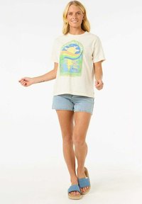 Camiseta color crema con un diseño gráfico colorido que presenta un sol y palmeras, combinada con pantalones cortos de mezclilla claros y sandalias azuladas tipo slip-on.