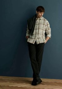 Pledota krekls smilškrāsas un zaļās nokrāsās, melni bikses un melnas kurpes. Tumšs džemperis pārmetts pāri plecam; koka grīda, zila siena fons.