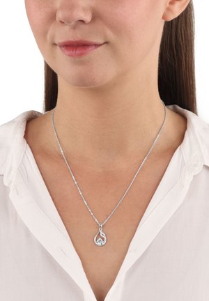 Collana d'argento con pendente a forma di goccia che presenta una pietra preziosa trasparente e dettagli intricati, indossata su una camicia bianca.