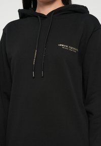Čierna mikina s kapucňou vyrobená z mäkkej látky, obsahujúca šnúrku na kapucni. Zlatý textový logotyp na ľavej strane hrude znie "ARMANI EXCHANGE MILANO/NEW YORK."