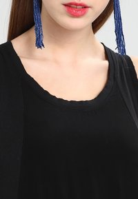 Czarna bezrękawnik z okrągłym dekoltem, o gładkiej fakturze. Długie niebieskie kolczyki w kształcie frędzli, które dodają kontrastu kolorystycznego.