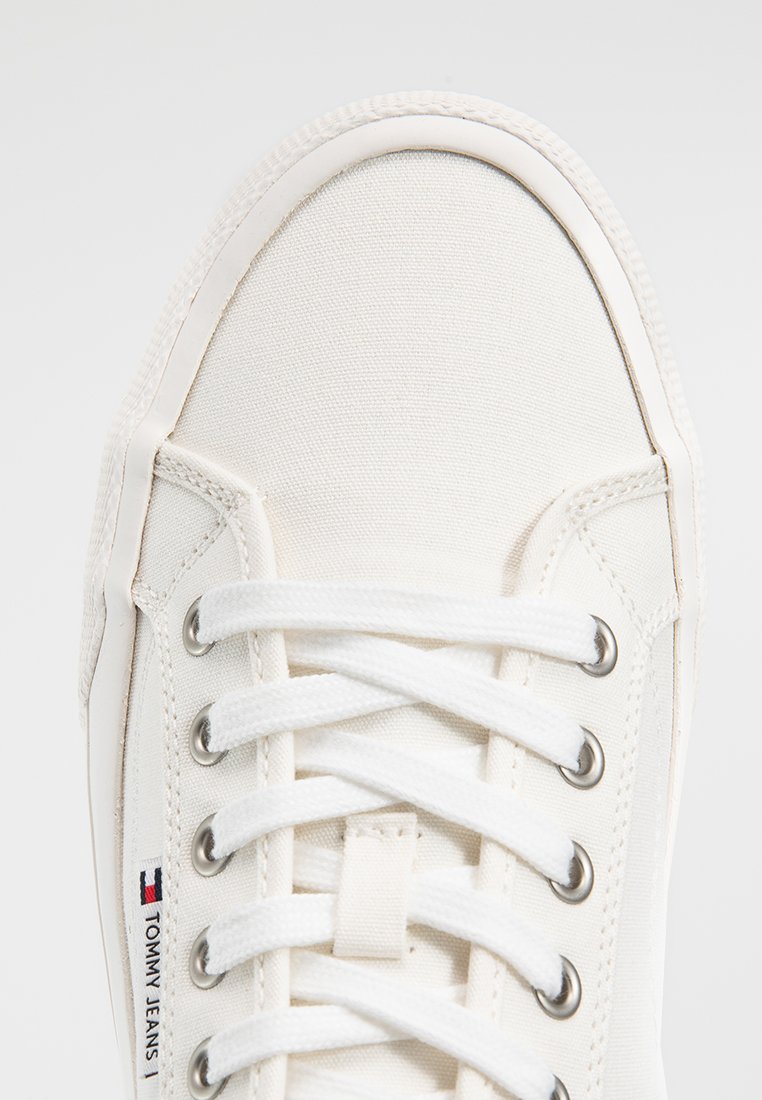 Zapatilla de lienzo blanca con cordones blancos, ojales de metal, suela de goma y etiqueta con el logo "Tommy Jeans" en el costado.