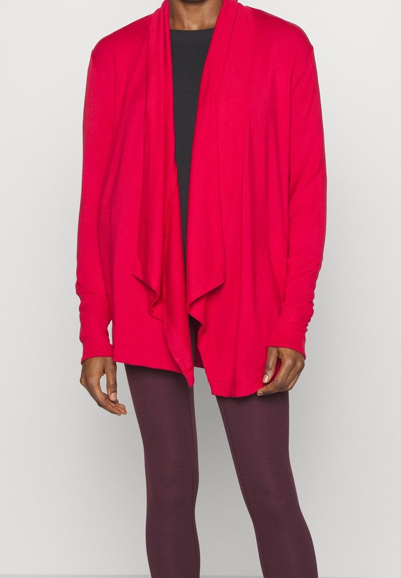 Cardigan long à manches rouges avec drapé devant ouvert, en tissu léger, porté avec un top noir et des leggings bordeaux. Texture lisse, coupe décontractée.