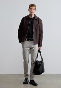 Hnedá textúrovaná bunda, čierna košeľa, svetlosivé nohavice, čierny opasok, čierna taška, čierne topánky; stojí pred svetlosivým pozadím.