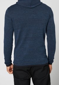 Dunkelblaue Kapuzenjacke mit strukturiertem Strick, ausgestattet mit einer großen Kapuze, eng anliegenden Ärmeln und einem Saum, der mit einem gerippten Abschluss versehen ist.