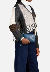 Gebreide cardigan met een mix van grijze, crèmekleurige en bruine texturen; heeft een V-hals, knoopsluiting en geometrische patronen. Houdt een blauwe leren clutch vast.
