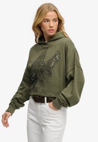 Felpa con cappuccio verde oliva con un grande disegno dettagliato di aquila in paillettes nere e argento, polsini a costine e vestibilità rilassata, abbinata a pantaloni bianchi.