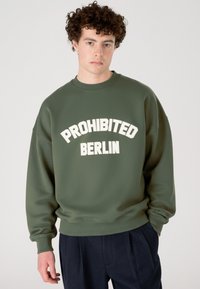 Jaunas vyras su garbanojais plaukais, vilkintis alyvuogių žalio spalvos džemperį su užrašu "PROHIBITED BERLIN" ir tamsiai mėlynas klostuotas kelnes, stovi prieš paprastą foną.