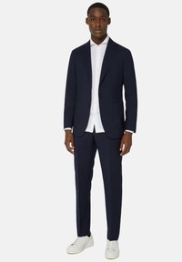 Mees, kes kannab tumesinist ülikonda, valget triiksärki ja valgeid sneakers'eid, seismas heledal ühtlasel taustal.