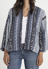 Veste de style kimono avec un motif géométrique bleu marine et blanc, dotée de larges manches et d'un design à l'avant ouvert. Fabriquée en tissu léger.