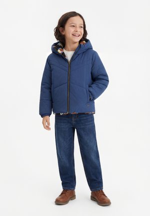 Niño sonriendo y de pie, vistiendo una chaqueta con capucha azul, vaqueros y botas marrones sobre un fondo blanco liso.