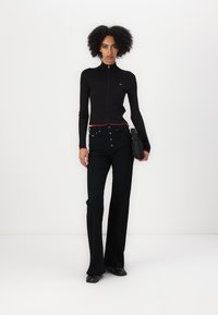 Svart, ribbet langermet genser med glidelås og rød detalj ved kragen, kombinert med høybuksete, svarte flared jeans og en svart håndveske.