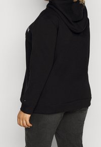 Person wearing a black hoodie with hood down and dark gray pants, standing sideways against a plain light background.