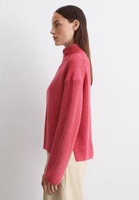 Rosa gestrickter Pullover mit hohem Kragen, gerippter Textur, seitlichen Schlitzen und langen Ärmeln. Über hellen Hosen getragen, schlicht gestylt.