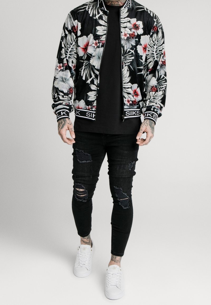 Černá bomber bunda s květinovým motivem bílých a červených květů, s žebrovanými manžetami a názvem značky na lemu. Kombinováno s černými roztrhanými úzkými džínami a bílými teniskami.
