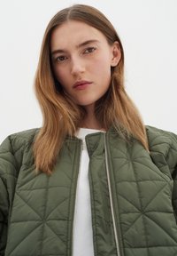 Zelená bomber bunda s prošíváním a zipovým uzávěrem, s geometrickým diamantovým vzorem a kulatým výstřihem, nošená přes bílý top.