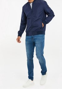 Marinblå bomberjacka med dragkedja, ribbade manschetter och fåll, i kombination med ljusblå jeans och vita sneakers, mot en enfärgad bakgrund.