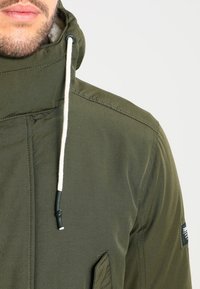 Oliivivihreä huppujakku, jossa on teksturoitu pinta, valkoiset nyörit, vino ompelu yksityiskohtina ja sivutasku.