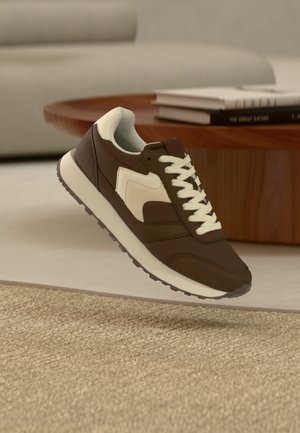 Brun og hvid sneaker med hvide snørebånd vist over et beige tæppe, nær et træ sofabord med stabel af bøger.