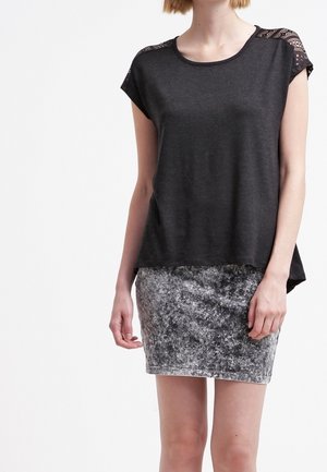 Femme portant un haut noir ample avec des épaules en dentelle et une mini-jupe grise texturée, se tenant devant un fond blanc uni.