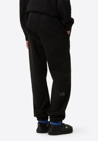 Pantalones jogger de forro polar negros con puños elásticos y bolsillos, con un logotipo discreto en el muslo. Llevados con calzado negro sin cordones y calcetines azules.