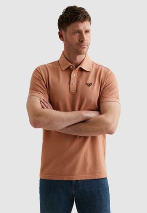 Mężczyzna z krótkimi włosami, noszący polo koloru łososiowego z wyhaftowanym logo, z założonymi rękami, patrzący w prawo.