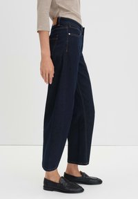 Jeans scuri, a vita alta, con gambe larghe, dettagli di cucitura sottili e una fettuccia di pelle marrone sulla cintura posteriore. Indossati con mocassini neri.