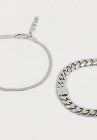 Δύο ασημένια βραχιόλια αλυσίδας σε λευκή επιφάνεια, το ένα με λεπτή επίπεδη αλυσίδα φίδι και το άλλο με χοντρές, στρογγυλεμένες κρίκους curb.