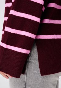 Bordeauxrode gestreepte trui met brede roze strepen, gebreide textuur en zijsplitten. Zichtbare manchetten en een ontspannen pasvorm op een lichtgrijze achtergrond.