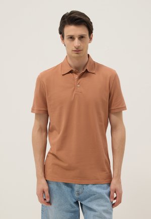 Jonge man staand, gekleed in een bruine korte mouwen poloshirt en lichtblauwe spijkerbroek, tegen een effen lichte achtergrond.