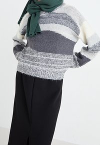 Pull en tricot à rayures horizontales gris, blanc et crème, avec une coupe décontractée. Associé à une jupe noire taille haute et une écharpe verte.