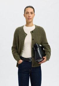 Olivgrön fluffig cardigan med rund hals och svarta knappstängningar, som bärs över en beige skjorta, tillsammans med mörk denim och en svart väska.