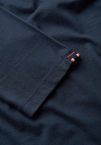 Camiseta de algodón azul marino con una textura suave, que presenta una costura visible y una pequeña etiqueta en naranja, blanco y azul marino en la manga.