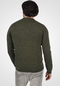 Olijfgroen gebreide trui met een gestructureerde afwerking. Voorzien van een ronde halslijn en ribgebreide manchetten. Gecombineerd met lichtgrijze jeans, die een casual pasvorm tonen.